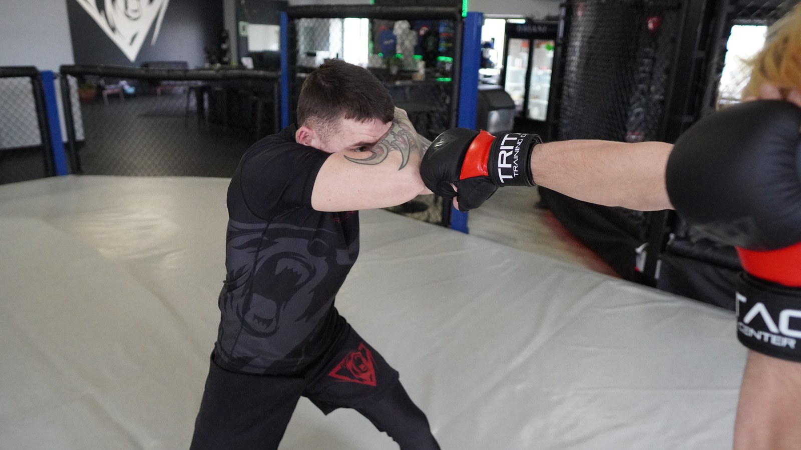A student throwing a straight punch at a training partner who covers up, on the mat beside the cage.