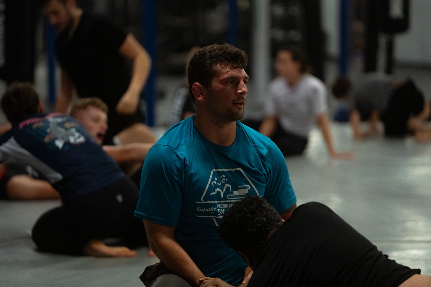 A coach kneeling on the mat correcting a student mid-drill while the class works around them.